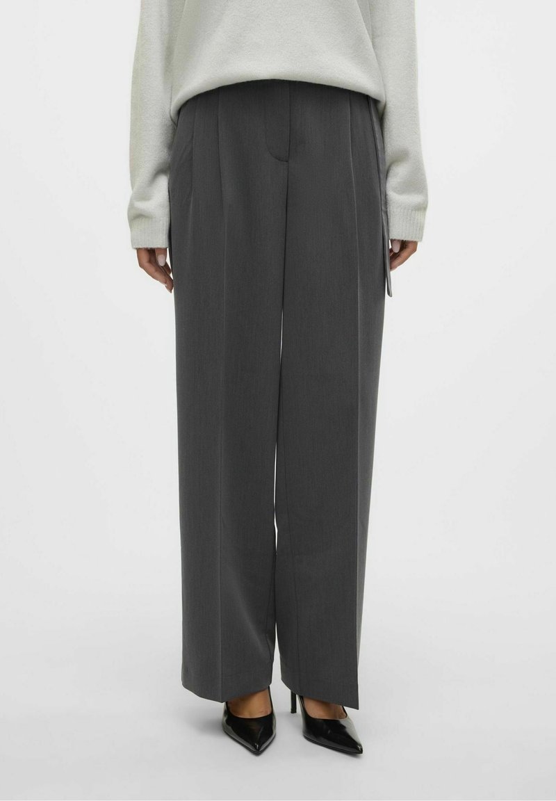 Personne portant un pantalon large gris foncé, un pull de couleur claire et des escarpins noirs à bout pointu, debout devant un fond uni.