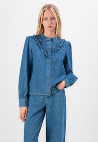 Denim shirt with ruffled collar, long sleeves, and button closure. Light blue color, soft texture, and visible stitching details.