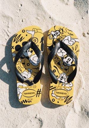 Világos sárga flip-flop papucsok fekete pántokkal, rajzfilmfigurák nyomataival, egymás mellett fekve texturált homokban.