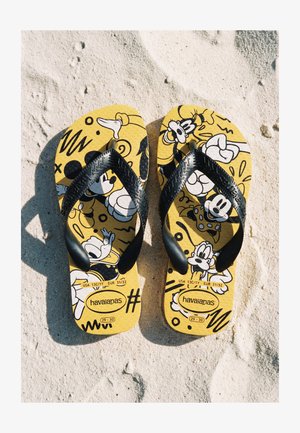 Tongs jaune vif avec des brides noires affichant des impressions de personnages de dessins animés, posées côte à côte sur du sable texturé.