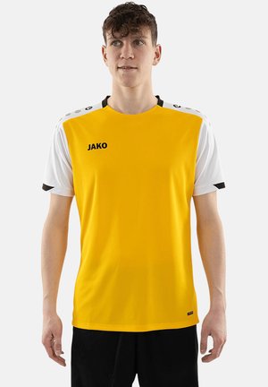 Giovane uomo che indossa una maglia sportiva gialla e bianca con pantaloncini neri, in piedi davanti a uno sfondo grigio chiaro.