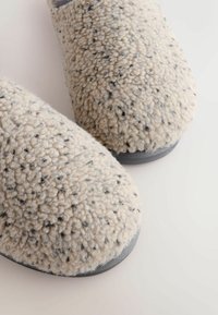 Soft, beige slipper with a textured, plush exterior featuring dark flecks. Rounded toe and gray rubber sole. Simple, cozy design.