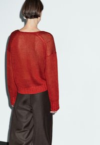 Maglione rosso lavorato a maglia, con maniche lunghe, vestibilità ampia e orlo a coste, abbinato a pantaloni a gamba larga marrone scuro. Dettaglio del filato texturizzato visibile.