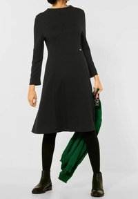 Svart långärmad klänning med utställd nederdel, tillverkad av mjukt tyg. Modellen bär svarta leggings och ankelstövlar, och håller i en grön tröja.