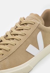 Beige suede sneaker with white rubber sole. Features include cotton laces and a prominent white logo on the side. Smooth texture and clean design.