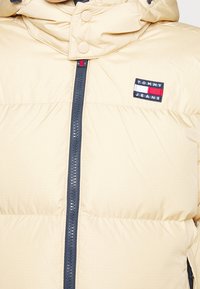 Veste matelassée beige avec fermeture éclair, accents bleu marine et un petit patch de marque avec "TOMMY JEANS" en plusieurs couleurs.