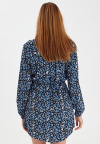Blaues Blumenkleid mit langen Ärmeln, gebundenem Bund und schwarzem Hintergrund. Es zeigt kleine Blumen in Blau und Grün aus leichtem Stoff.