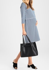 En gravid kvinna klädd i en blå och vit randig klänning bär en stor svart handväska och har på sig svarta öppna klackskor med blockklack.