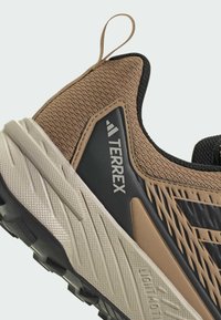 Brown textured hiking shoe with a mesh upper, black accents, and a chunky rubber sole. Features a pull tab and "TERREX" branding.