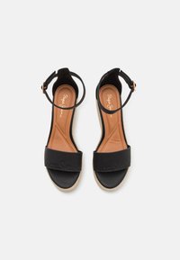 Svarte espadrillosandaler med bred reim, ankelspenne og jute-omviklet såle. Terskelen er polstret med en lys brunt læraktig tekstur.