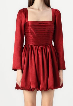 Kvinde iført en rød kjole med lange ærmer, firkantet udskæring, plisseret krop og en samlet, voluminøs nederdel.