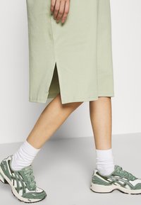 Jasnozielona sukienka midi z bocznym rozcięciem, noszona z zielono-białymi butami sportowymi i białymi skarpetkami sięgającymi do kostek. Gładka bawełniana tekstura.