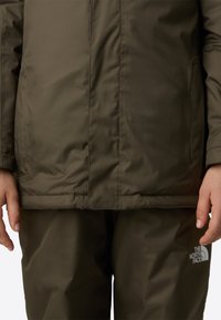 Olivengrønn isolasjonsjakke med horisontal søm og sydd kant. The North Face-logoen er synlig på venstre bryst og bukseben.