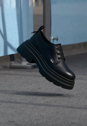 Black leather platform shoe with thick sole and black laces, suspended above pavement near a blue mesh fence and metal pole.
