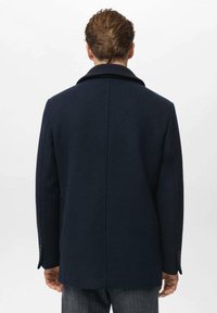Cappotto in lana blu navy con colletto ampio, taglio dritto e spalle strutturate. Presenta polsini con bottoni e un design pulito e minimalista.