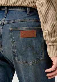 I jeans in denim blu scuro presentano una patch in pelle con il logo "Wrangler" e una cucitura a zigzag sulla tasca posteriore. È visibile la texture del maglione.