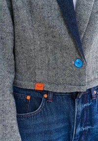 Detail sivého blazeru so vzorom rybej kosti s jasne modrým gombíkom, doplnený tmavomodrými džínsami s oranžovými nitmi a malou oranžovou značkou.