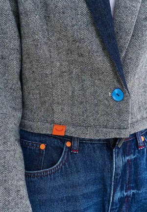 Zbliżenie na szarą marynarkę w jodełkę z jasnoniebieskim guzikiem, zestawioną z ciemnoniebieskimi dżinsami z pomarańczowymi nitami i małą pomarańczową metką.