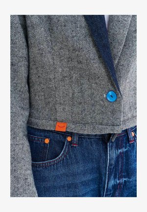 Zbliżenie na szarą marynarkę w jodełkę z jasnoniebieskim guzikiem, zestawioną z ciemnoniebieskimi dżinsami z pomarańczowymi nitami i małą pomarańczową metką.