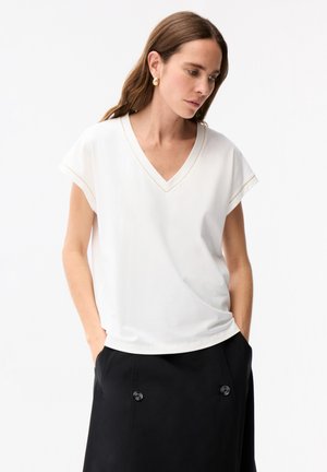 Kvinde med langt brunt hår iført guldringe i ørerne, en hvid V-udskæring med korte ærmer og et sort skørt med store knapper, hænderne i lommerne.