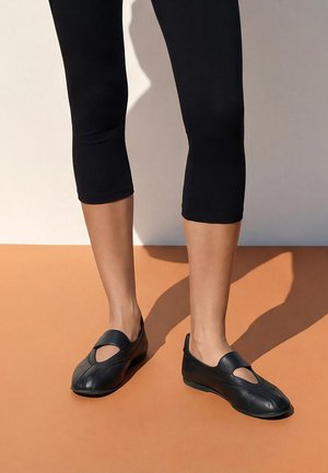 Legs wearing black cropped leggings and black closed-toe flats with a round cutout on top, standing on a two-tone floor.