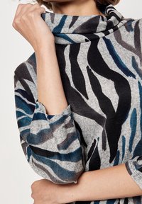 Haut en maille grise avec un col bénitier aux rayures zébrées bleues et noires, manches longues avec revers roulés, et tissu doux et texturé.