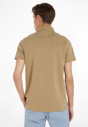 Muž oblečený v béžovom polo tričku s krátkym rukávom a svetlomodrých nohaviciach stojí chrbtom ku kamere.