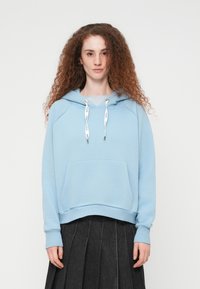 Νεαρή γυναίκα με σγουρά κόκκινα μαλλιά που φοράει ένα ανοιχτό μπλε φούτερ με λευκά κορδόνια και μια σκούρα πλισέ φούστα, στέκεται μπροστά σε ένα λευκό φόντο.