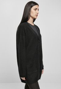 Black cardigan made of textured fabric, featuring a V-neck, two buttons, and a front pocket. Long sleeves extend to the wrists.