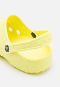 Sabates tipo clogs amarelas Crocs com uma superfície texturizada, parte traseira aberta e grandes orifícios laterais. Apresentam um logótipo circular da marca na alça. Design leve.