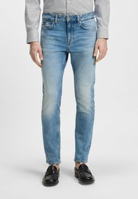 Jeans en denim bleu clair avec une coupe fuselée, présentant une texture décolorée, un design à cinq poches et une fermeture à bouton. Portés avec des mocassins noirs.