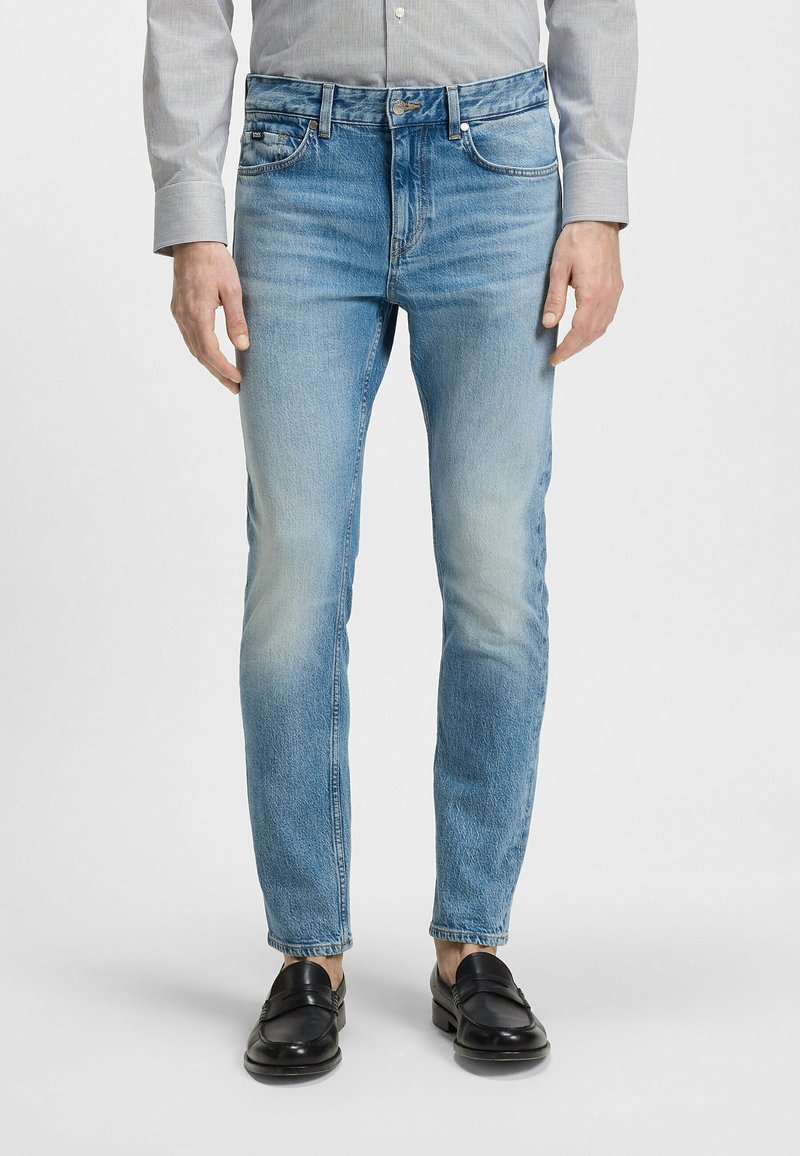 Jeans en denim bleu clair avec une coupe fuselée, présentant une texture décolorée, un design à cinq poches et une fermeture à bouton. Portés avec des mocassins noirs.