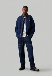 Set iz temno modre denim jakne in hlač. Jakna ima logo in elastični pas. Hlače so s žepi in ravnim krojem, v kombinaciji s črnimi čevlji.