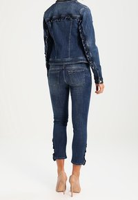 Jeansjacka med volangdetaljer längs ärmarna, tillsammans med ankellånga skinnyjeans, båda i mörkblått. Accentuerad med knappdetaljer.