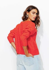 Pull en tricot rouge avec un motif en losange, des poignets côtelés et un détail au dos. Associé à un jean taille haute bleu clair.