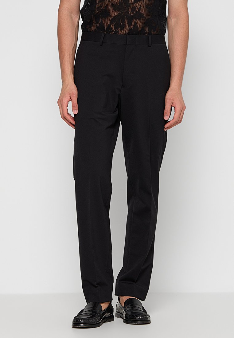 Una persona sta in piedi indossando pantaloni neri e una camicia nera trasparente e decorata, abbinata a mocassini neri, su uno sfondo bianco semplice.