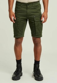 Pantaloni cargo verde oliva con molte tasche, dettagli con zip, risvolti e un design aderente, abbinati a stivali neri con lacci.