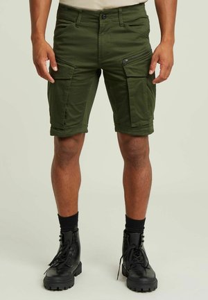 Pantaloni cargo verde oliva con molte tasche, dettagli con zip, risvolti e un design aderente, abbinati a stivali neri con lacci.