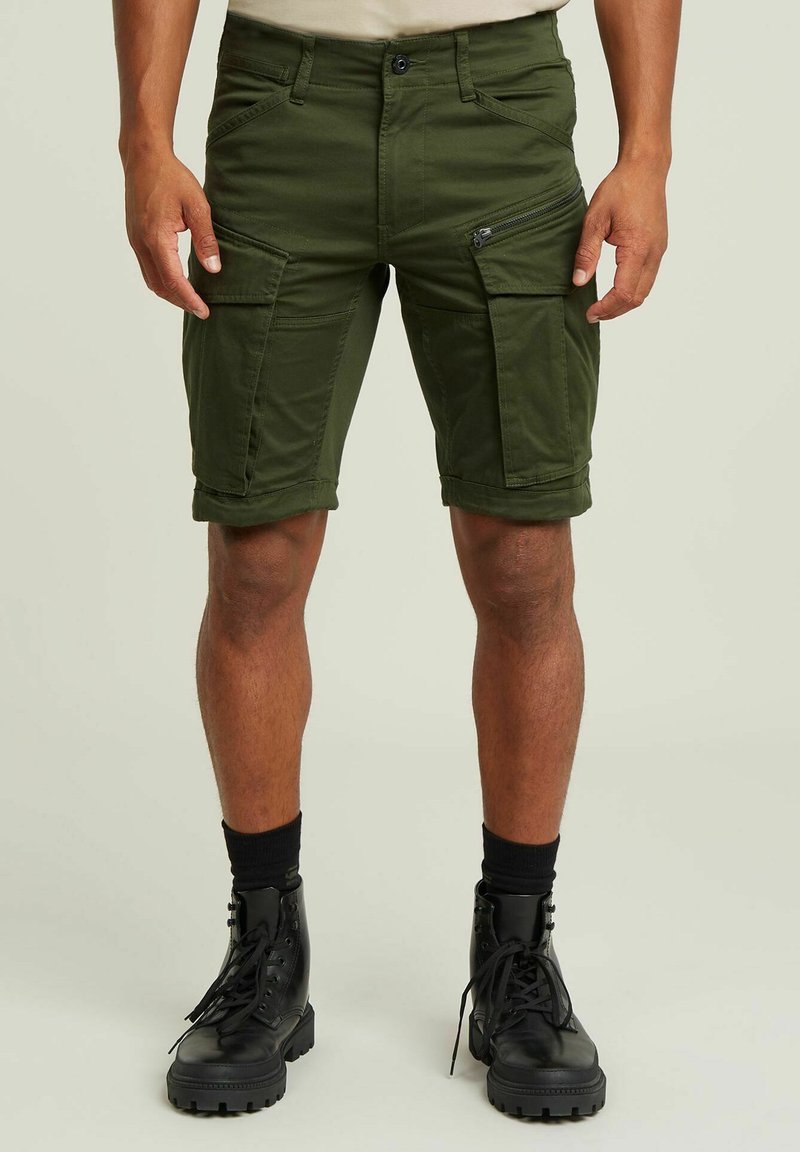 Pantaloni cargo verde oliva con molte tasche, dettagli con zip, risvolti e un design aderente, abbinati a stivali neri con lacci.