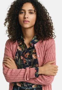 Rosa Strickjacke mit Kabelmuster und gerippten Kanten, kombiniert mit einem floral bedruckten Hemd mit dunklem Hintergrund.