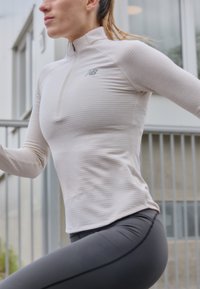 Haut de sport beige clair avec un col montant, des rayures texturées et une fermeture éclair quart, associé à un pantalon noir ajusté. Détails du logo réfléchissant.