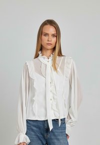 Weiße Bluse mit Rüschendetails, hohem Kragen und transparenten langen Ärmeln. Verfügt über eine Knopfleiste auf der Vorderseite und ein gebundenes Halstuch. Getragen mit blauen Jeans.