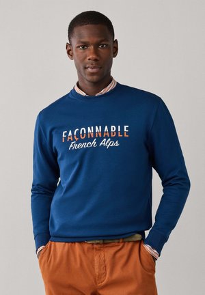 Homme portant un sweat-shirt bleu avec le texte "Façonnable French Alps" et un pantalon orange, debout les mains dans les poches sur fond uni.