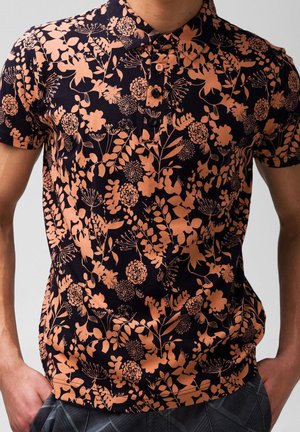 Tummansininen poolopaita, jossa on persikanvärisiä kukka- ja lehtikuvioita, kauluksessa kolme nappia, yllään henkilö, jonka kädet ovat taskuissa.