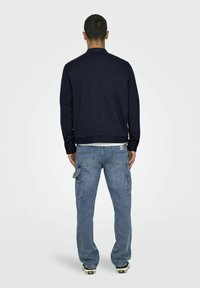 Marineblå casual jakke med høy krage, parret med lyseblå cargo-jeans med sidelommer og oppbrettede jakkesømmer, iført med svarte joggesko.