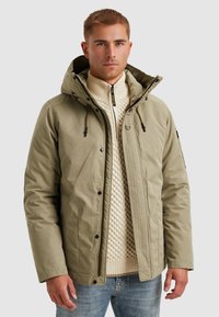 Khaki pufferväst med huva, med dragkedja, elastiska dragsnören och en subtil textur. Bärs över en krämfärgad stickad tröja.