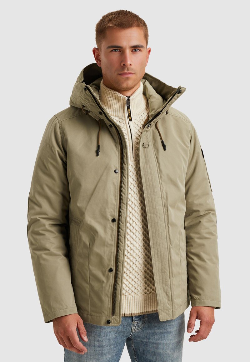 Khaki pufferväst med huva, med dragkedja, elastiska dragsnören och en subtil textur. Bärs över en krämfärgad stickad tröja.