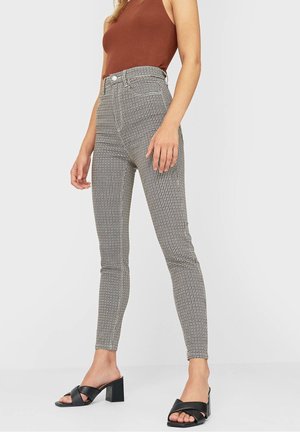 Kobieta w wyszczuplających, wysokim stanem spodniach w kratę, bezrękawniku w kolorze brązowym oraz czarnych sandałach na słupku z odkrytymi palcami, stojąca na białym tle.