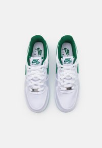 White Nike Air Force 1 sneakers with green accents, featuring a smooth upper, round toe, and a silver logo plate on the lacing.
