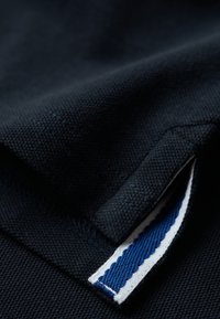 Navy katoen stof met een gestructureerde weving; bevat een decoratief label met blauwe en witte strepen aan de rand. Close-up van een gevouwen kledingstuk.